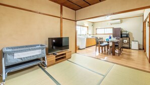 A traditional Japanese-style room with tatami flooring, a television on a wooden stand, and a kitchen area visible through an open doorway.