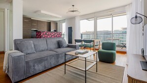 Ein modernes Wohnzimmer mit einem grauen Sofa, einem kleinen Tisch und einem grünen Sessel. Ein Küchenbereich mit Holzschränken und einer roten, gemusterten Rückwand. Das Zimmer verfügt über große Fenster mit Blick auf die Stadt.