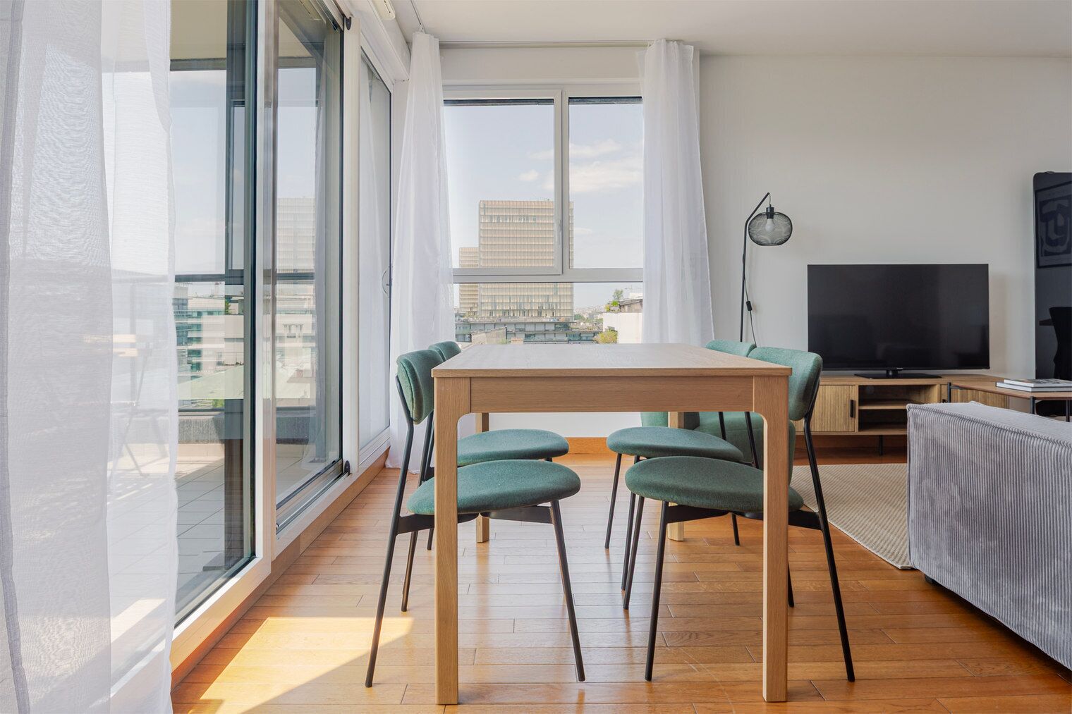 A modern living room with a wooden dining table, green upholstered chairs, a flat-screen TV, and a large window offering a city view.