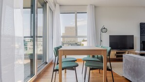 Ein modernes Wohnzimmer mit einem Holzesstisch, grünen Polsterstühlen, einem Flachbildfernseher und einem großen Fenster mit Blick auf die Stadt.
