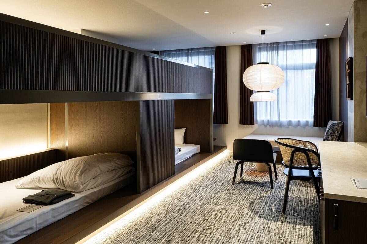A modern hotel room with bunk beds, a desk, chairs, and a large window with curtains.
