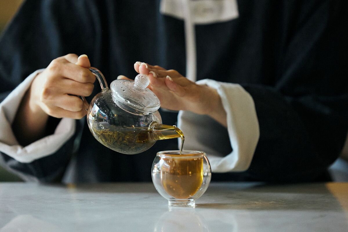 Eine Person gießt Tee aus einer Glas-Teekanne in eine Tasse.