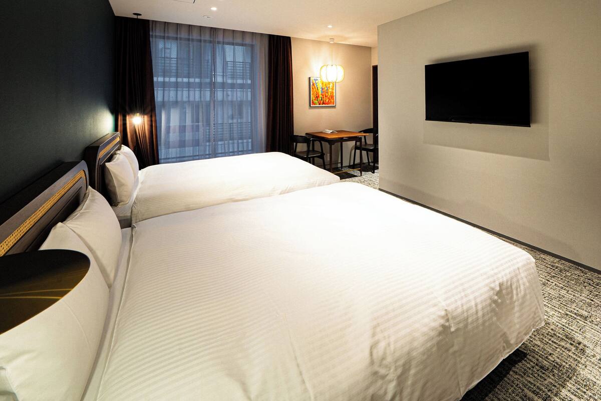 A hotel room with two beds, a desk, a chair, a TV, and a large window with curtains.