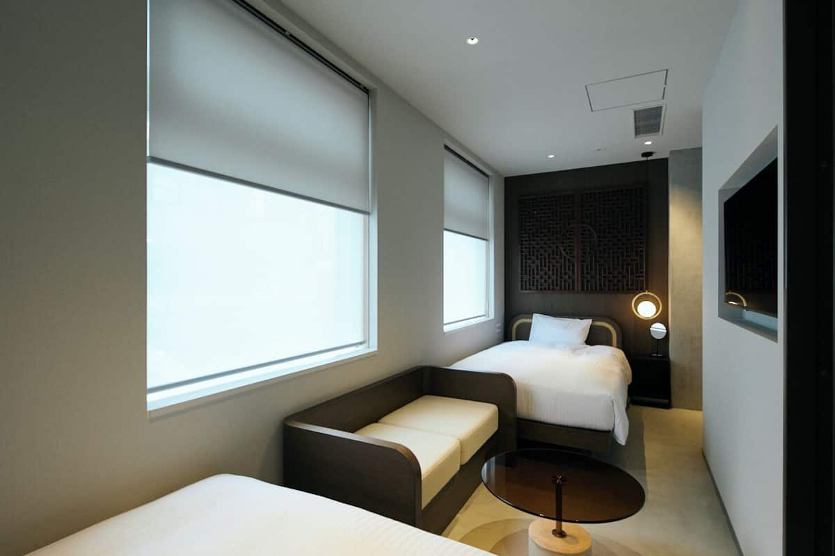 A modern hotel room with two beds, a sofa, a small table, and large windows.