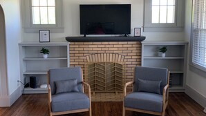 A living room with a brick fireplace, two grey armchairs, a flat-screen TV mounted above the fireplace, and white shelving units on either side. - Renovated In-Town Bungalow in French Lick (French Lick)