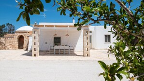 Un bâtiment blanc avec une entrée en forme d’arc en pierre, un espace extérieur abrité avec des sièges, et une structure en forme de dôme en arrière-plan.