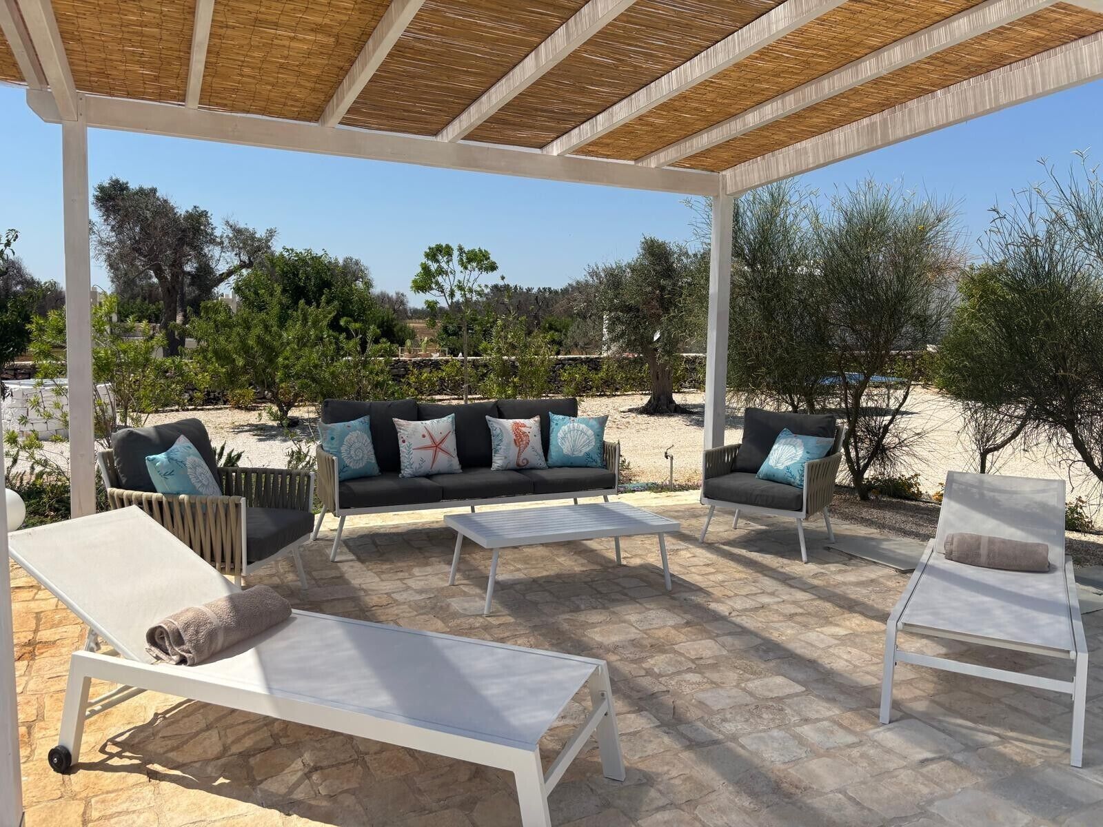 Un patio exterior con zona de estar cubierta, suelo de piedra y abundante vegetación.
