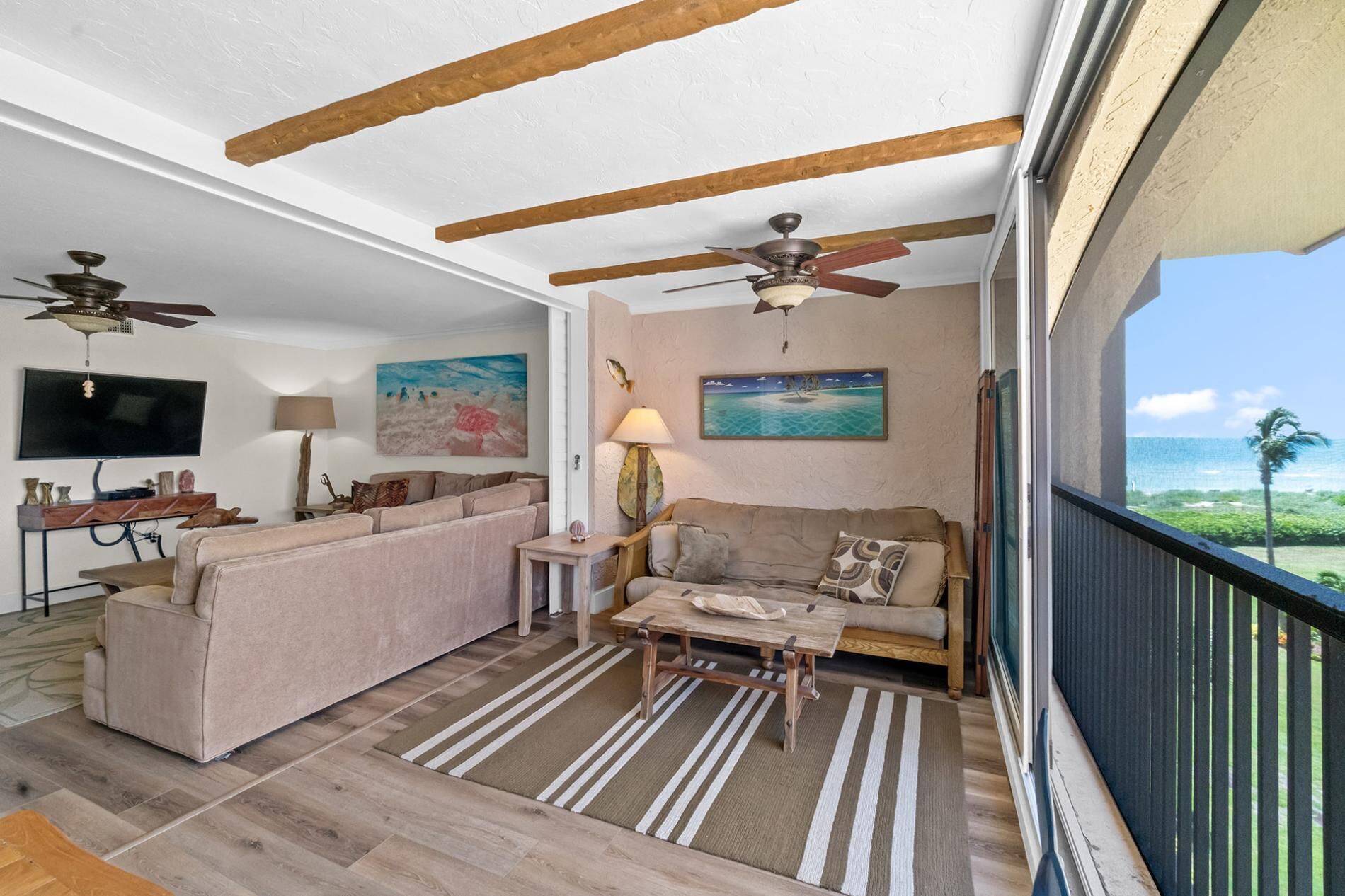 A spacious living room with a large sofa, a coffee table, and a TV. There are ceiling fans, a painting, and a view of the ocean through a balcony.