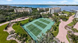 A large tennis court surrounded by buildings, a body of water, and a clear sky.