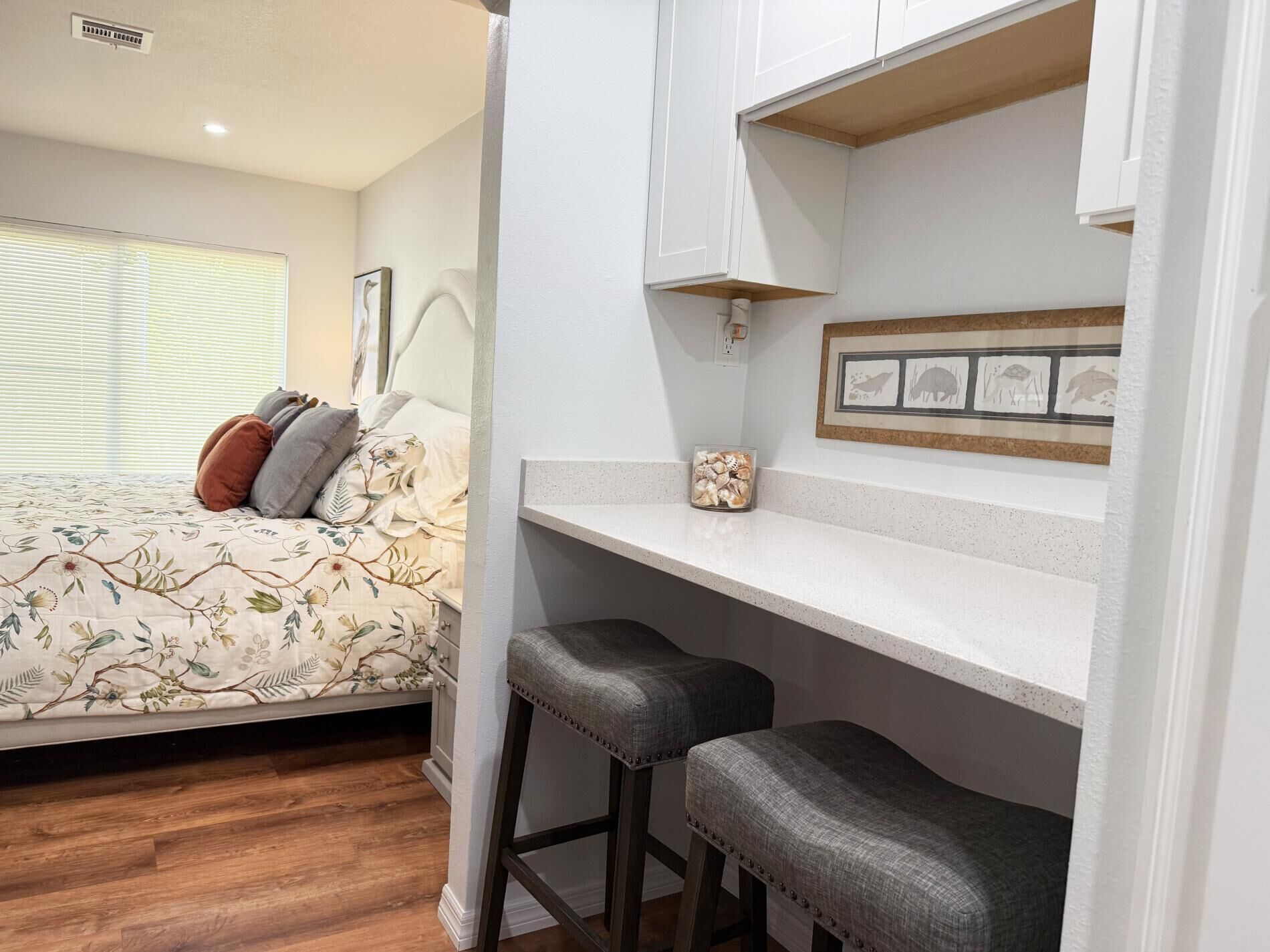 Une chambre à coucher comprenant un lit, une table de chevet, un petit bureau avec deux tabourets, une fenêtre avec des stores et une étagère murale agrémentée d’objets décoratifs.