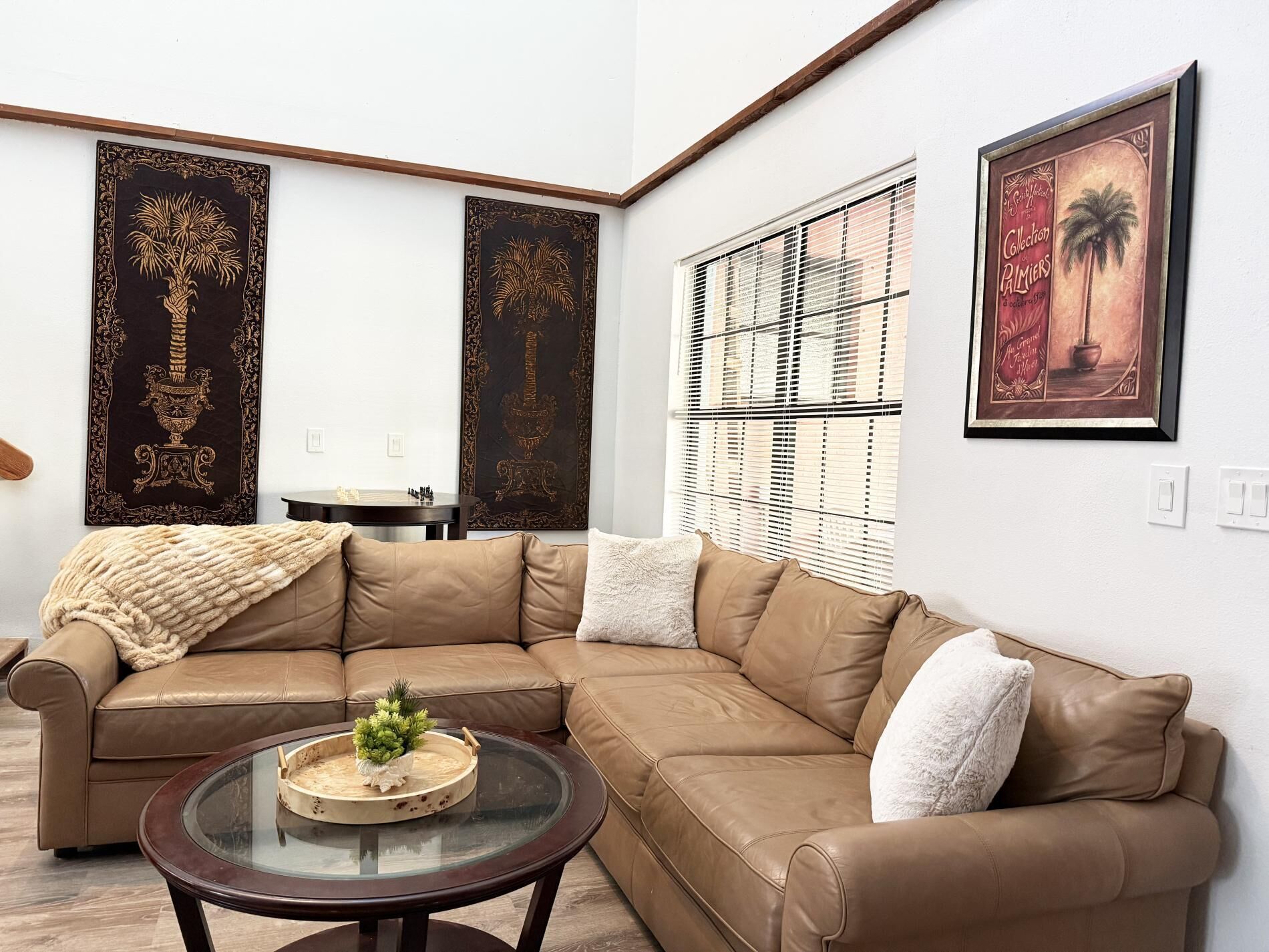 Una sala de estar con un sofá de cuero marrón, una mesa de centro de vidrio con una planta y dos paneles decorativos en la pared.