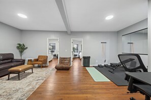 Una sala moderna con un sofá de cuero marrón, un sillón marrón, una mesita redonda y un banco. En el suelo, hay una esterilla de yoga y un juego de pesas.
