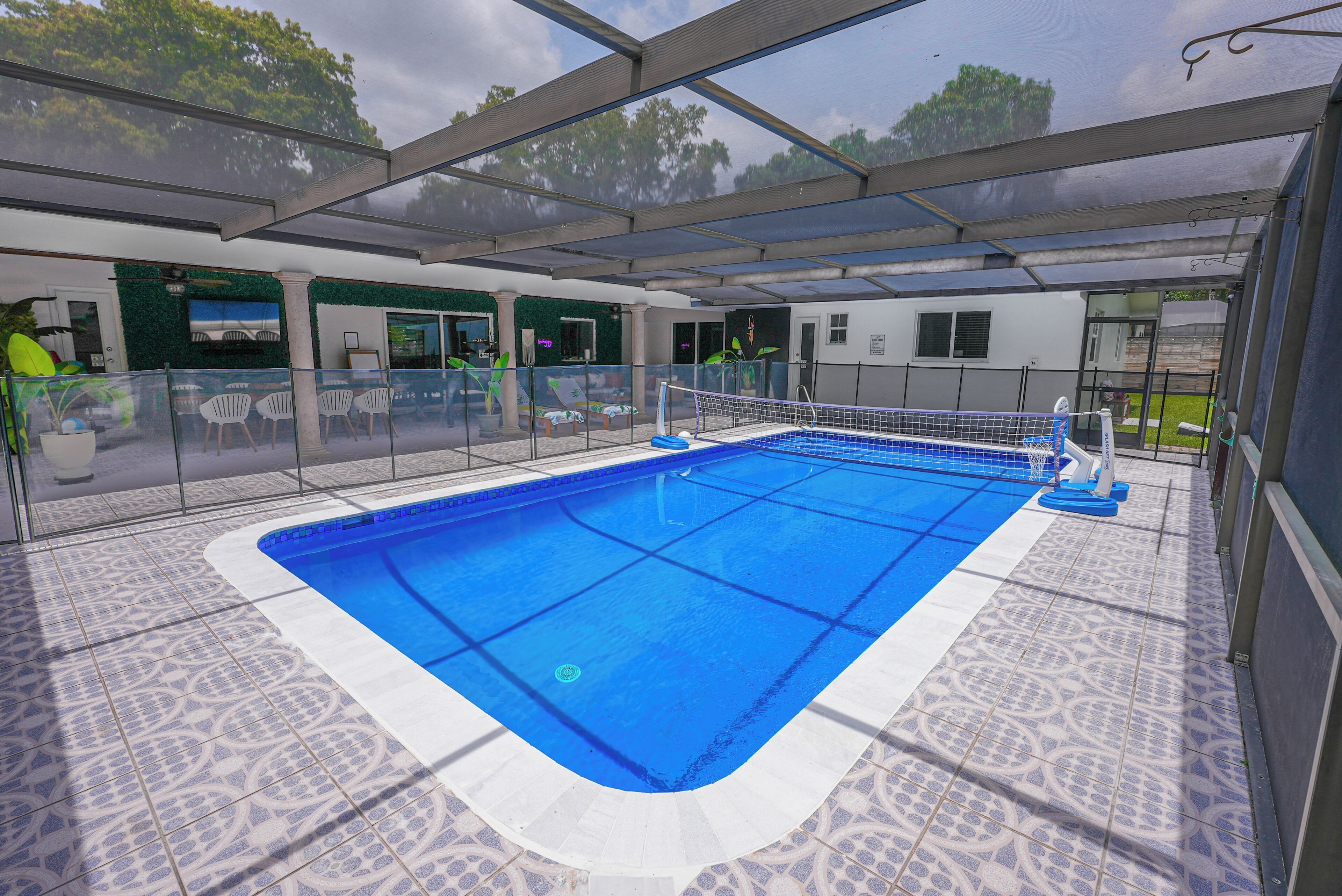 A covered swimming pool with a glass barrier, surrounded by patterned tiles, and a view of the outdoors.