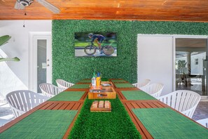 Uma área de jantar com uma mesa de madeira comprida, cadeiras brancas e um centro de mesa com grama sintética verde.