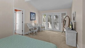 Une chambre à coucher comprenant un lit, deux fauteuils à bascule, une petite table avec une lampe, un miroir et une porte-fenêtre coulissante donnant sur un balcon.