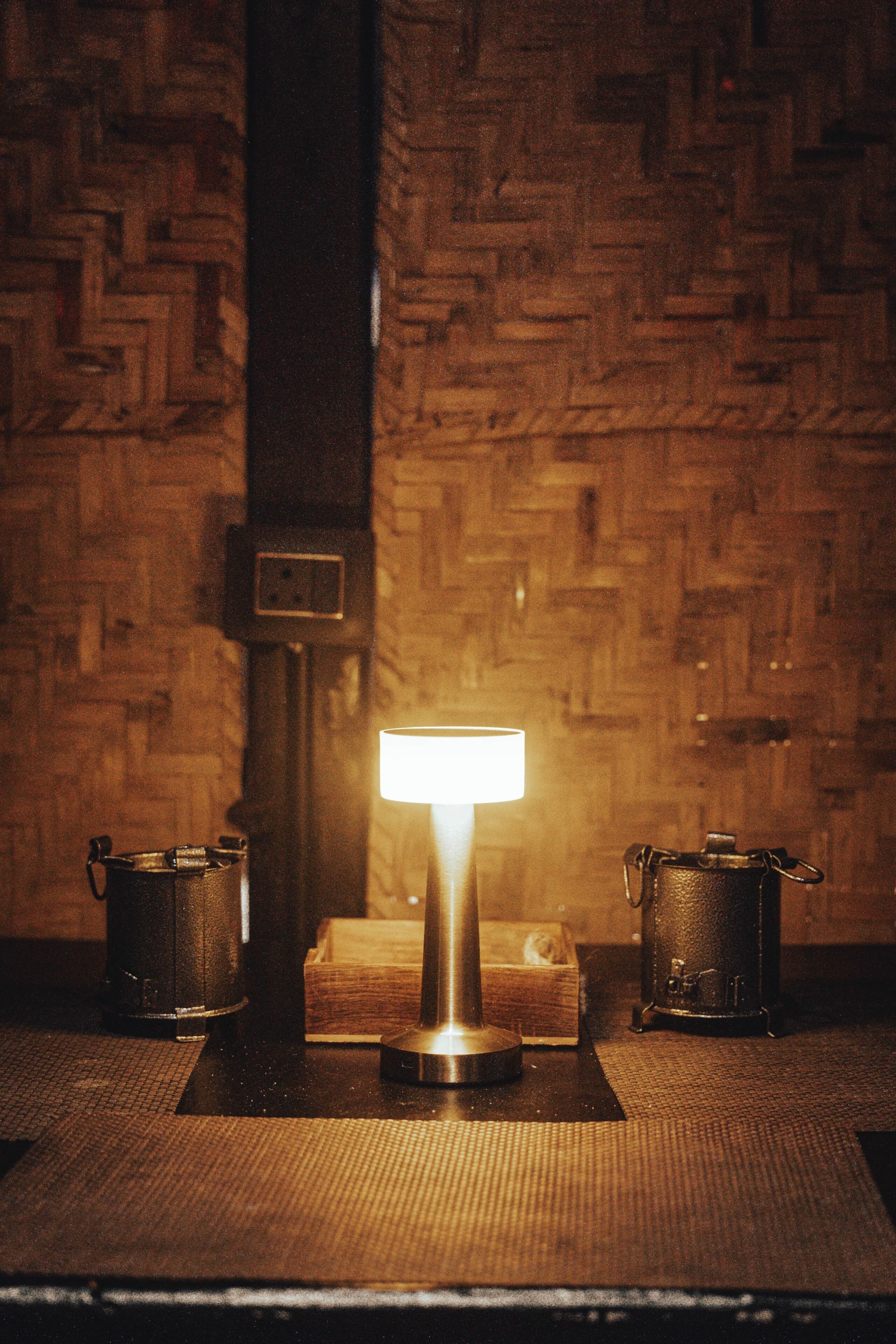 A table with a lamp, two metal containers, and a wooden box against a brick wall.