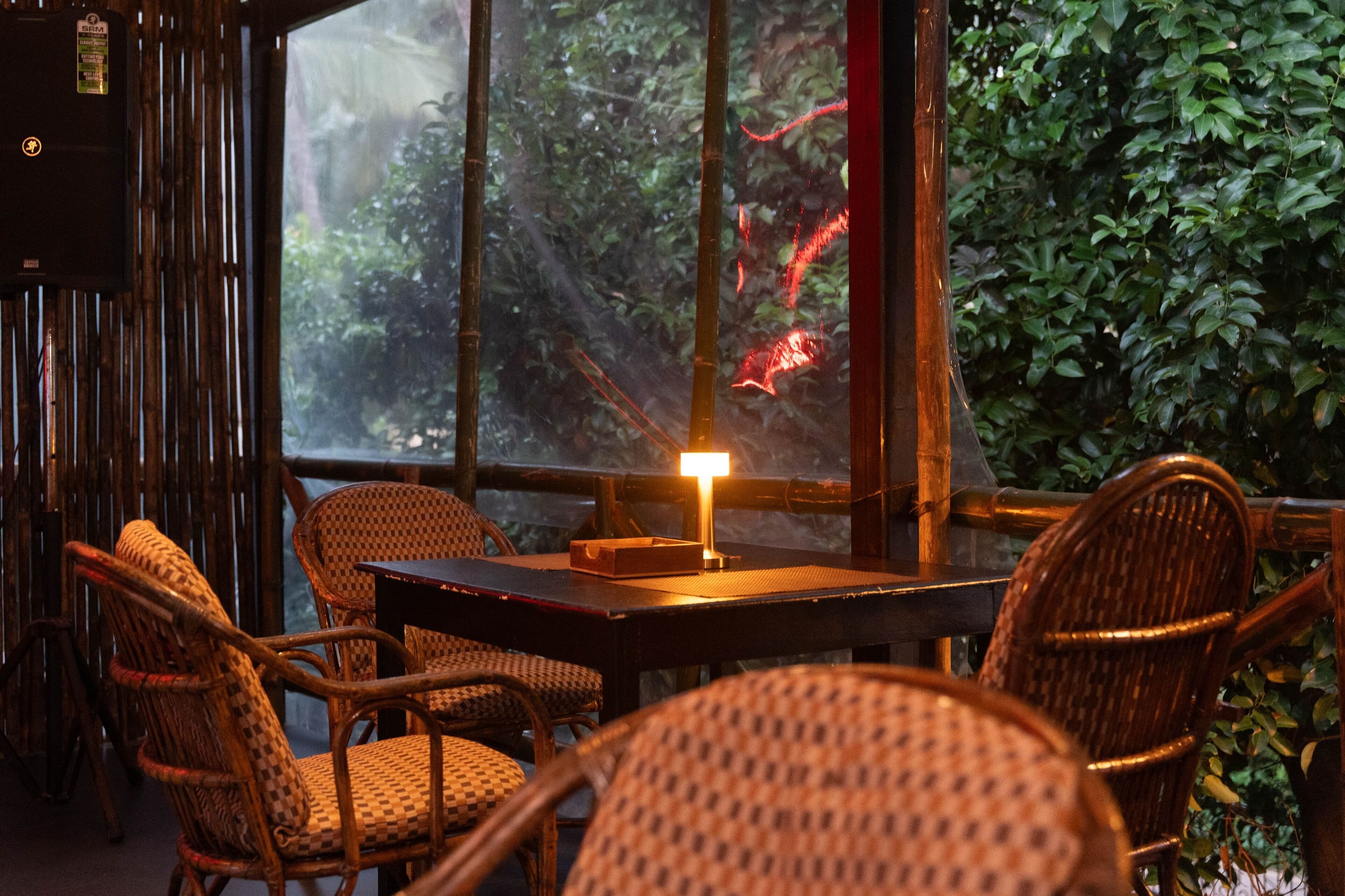 Un restaurante con mobiliario de mimbre, una mesa con una lámpara encendida y vistas a la vegetación a través de amplias ventanas.