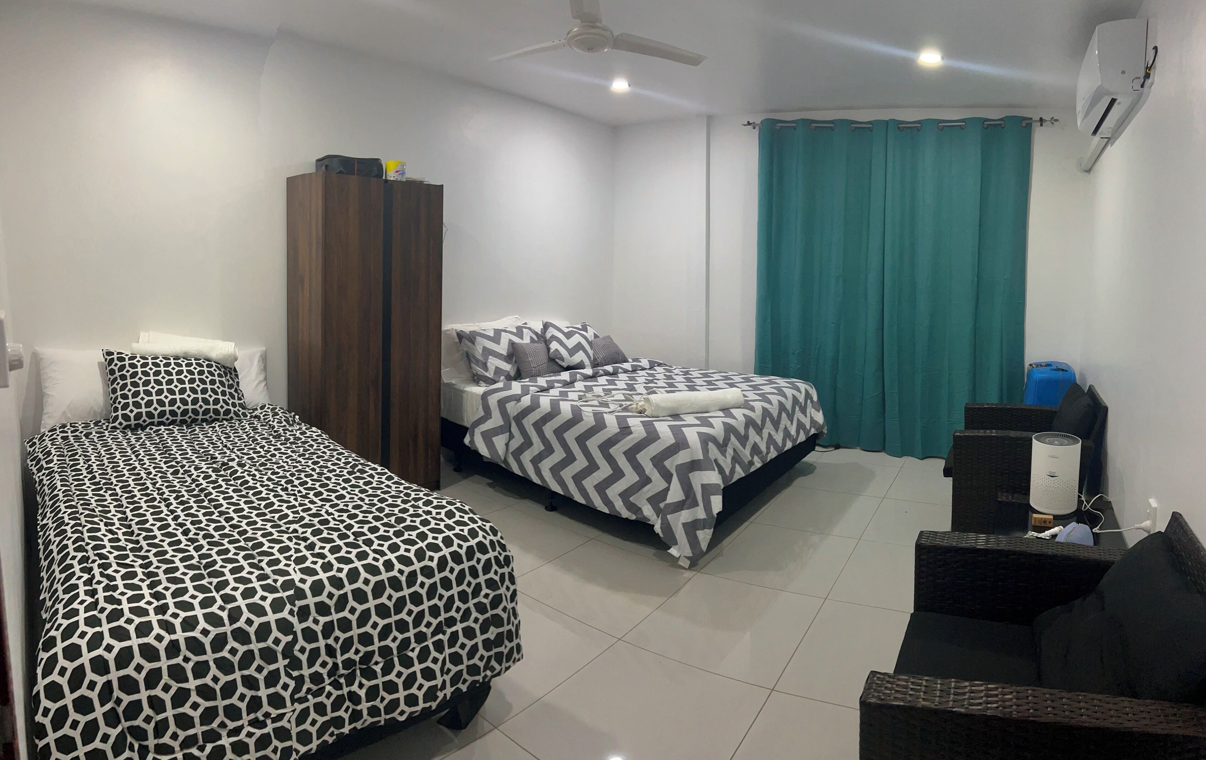 A hotel room with two beds, a wardrobe, a ceiling fan, an air conditioner, a teal curtain, and a small seating area with a chair and a table.
