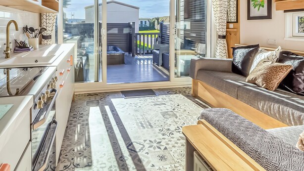 A modern kitchen with a built-in oven, a dining area with a sofa and patterned cushions, and a sliding glass door leading to an outdoor patio. - Pladda Pod with Hot Tub S (Ayr)