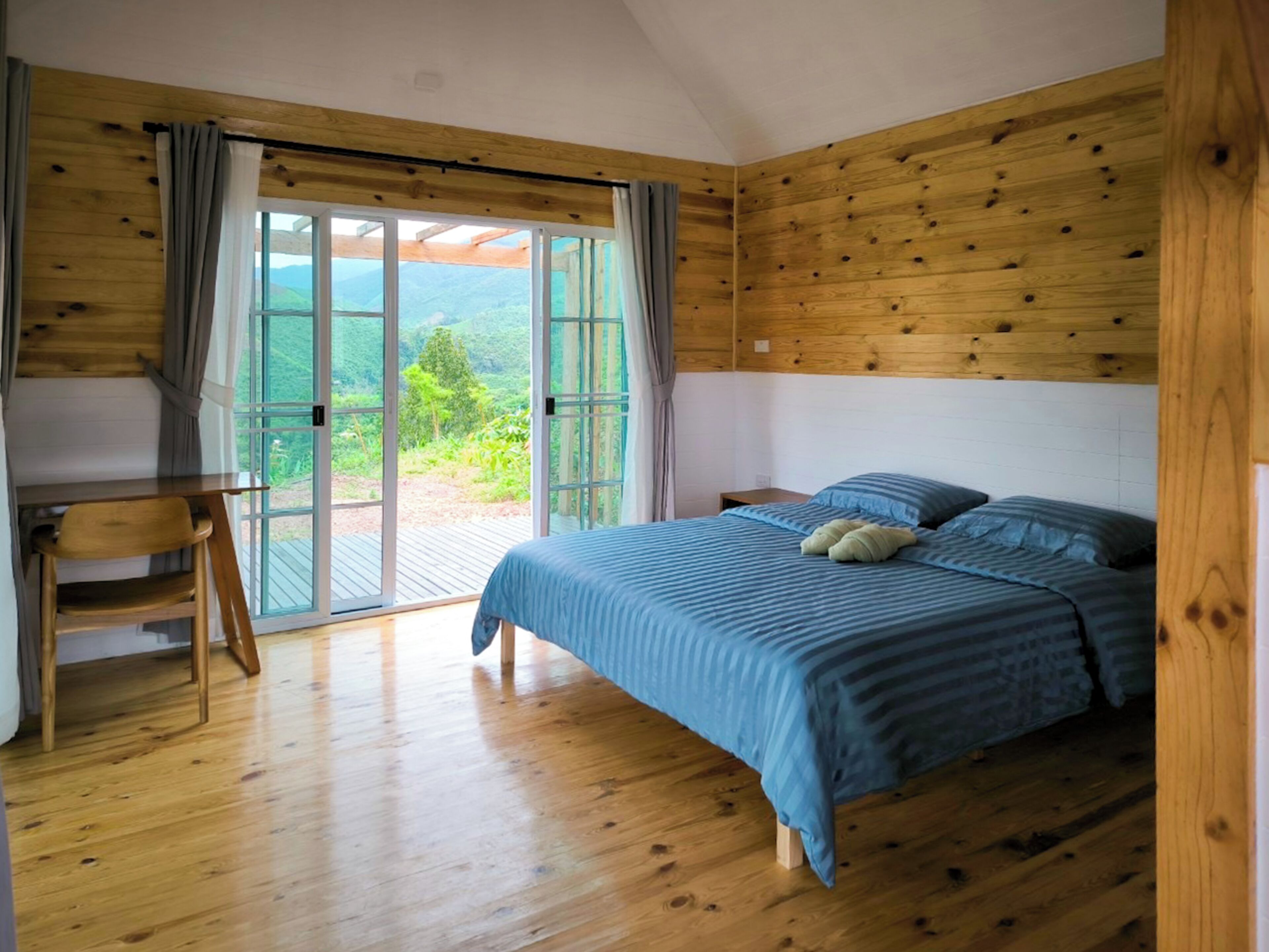 Una camera da letto con pavimento in legno, un letto con biancheria azzurra, una scrivania con sedia e un'ampia porta scorrevole in vetro che dà su un balcone con vista sul verde.