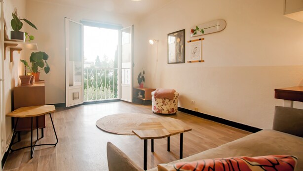 Una sala de estar moderna con piso de madera, un tapete redondo, un sofá con cojín estampado, una mesita de madera y una silla. Hay plantas en macetas en un estante y una puerta que da a un balcón exterior.