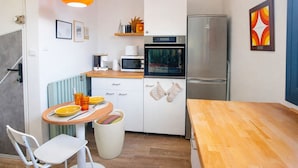 Cocina compacta con microondas, horno y refrigerador. Hay una mesa de comedor con sillas y un cubo de basura pequeño.