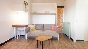 Una sala de estar con un sofá, una mesita de madera, una silla blanca, un radiador y una planta en un estante.