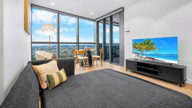 A modern living room with a large flat-screen TV, a sofa, a dining table with chairs, and a view of the city through large windows.