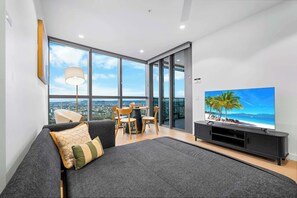 Una sala moderna con un televisor de pantalla plana grande, un sofá, una mesa de comedor con sillas y vistas a la ciudad a través de grandes ventanales.