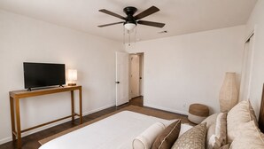 Ein Hotelzimmer mit einem Bett, einem Fernseher auf einer Holzkonsole, einem Deckenventilator und einer Tür zu einem anderen Raum.