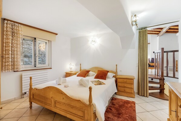 Ein Schlafzimmer mit einem hölzernen Bett, Nachttischen, einem Beistelltisch, einem Fenster mit Vorhängen, einer Chandelüster und einer Treppe.