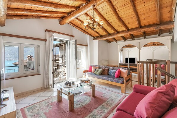 A spacious living area with a wooden ceiling, a sofa set, a glass coffee table, and a view of mountains through a large window.