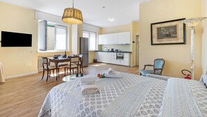 A bedroom with a bed, a dining table with chairs, a kitchen area, and a TV mounted on the wall.