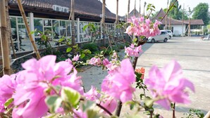 Un edificio con techo de tejas, zona de estacionamiento y flores rosadas en primer plano.