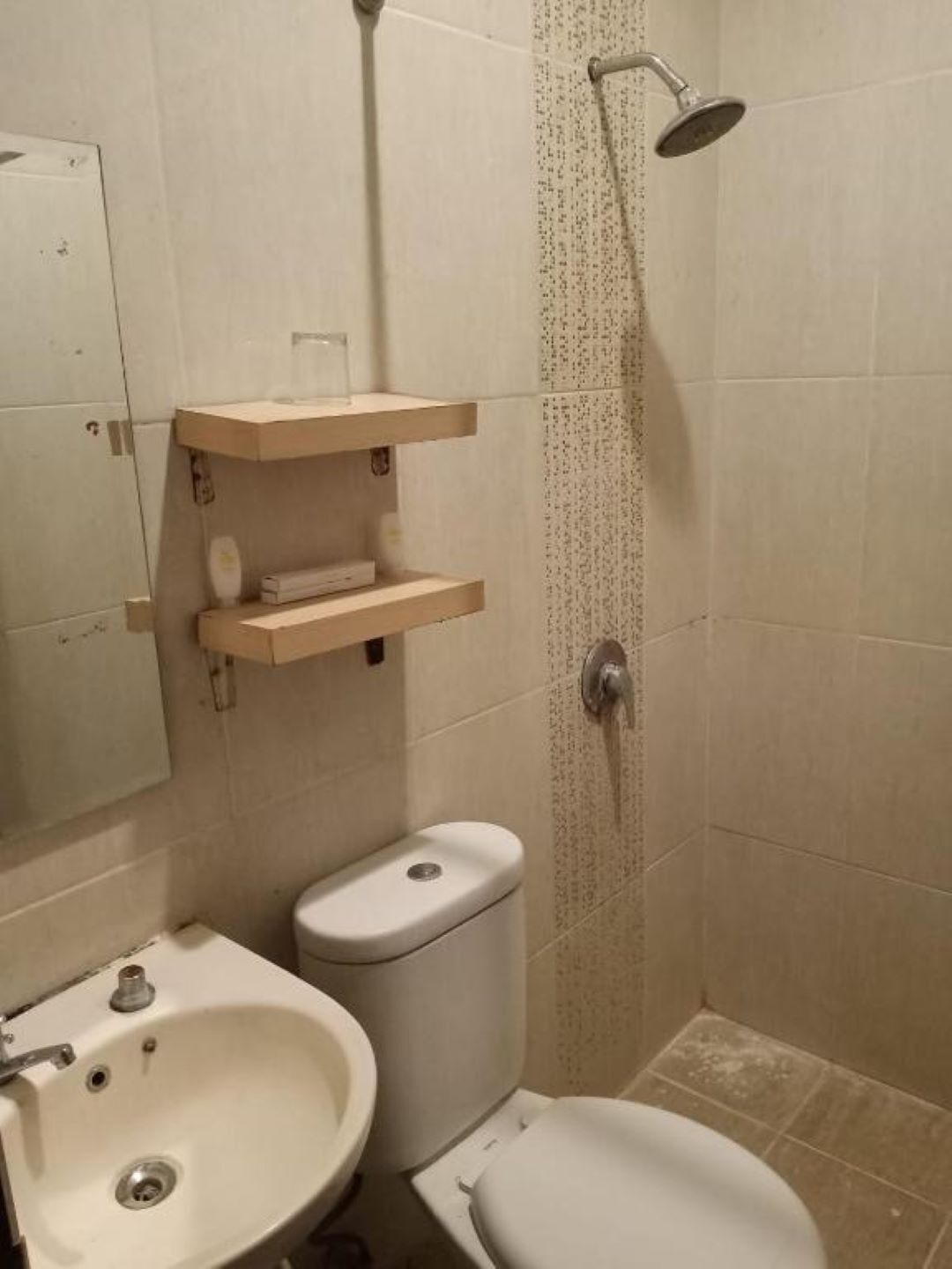 A bathroom with a toilet, sink, and shower. There are shelves above the toilet and a mirror on the wall.
