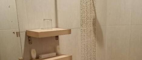 A bathroom with a toilet, sink, and shower. There are shelves above the toilet and a mirror on the wall.