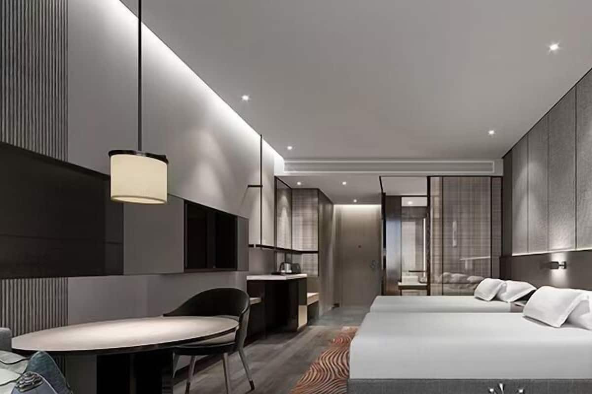 A modern hotel room with a large bed, a desk, and a chair. There is a window with curtains, a wall-mounted TV, and a ceiling light.