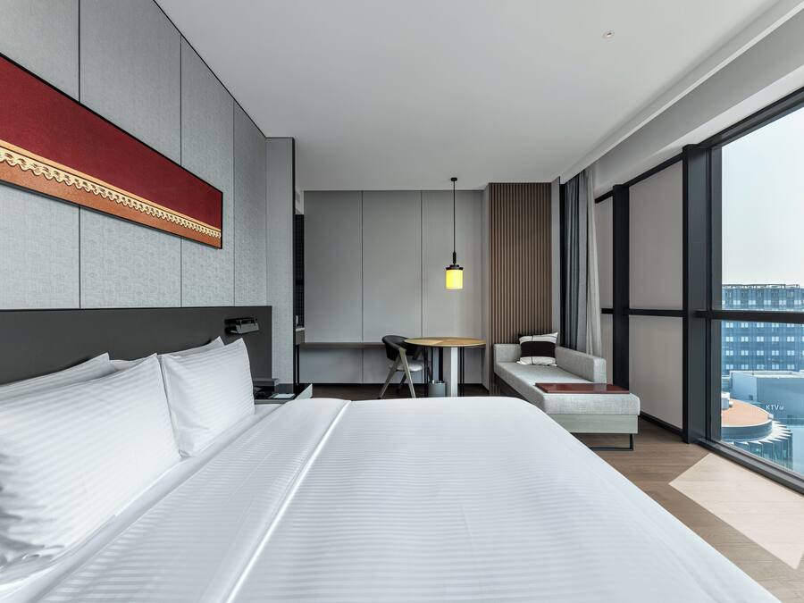 Una habitación de hotel moderna con una cama grande, un escritorio, una silla y un sofá. Hay un ventanal con vistas a la ciudad.