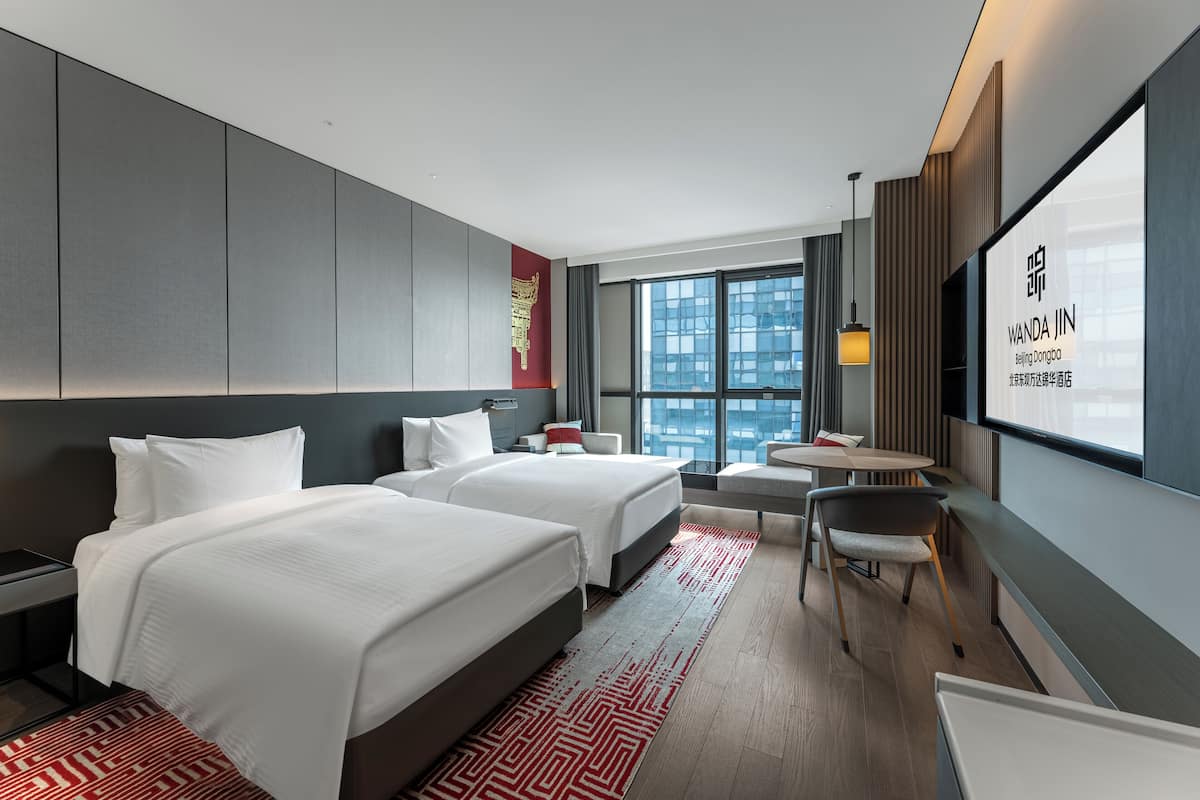 A modern hotel room with two beds, a small table, a chair, and a TV on the wall.