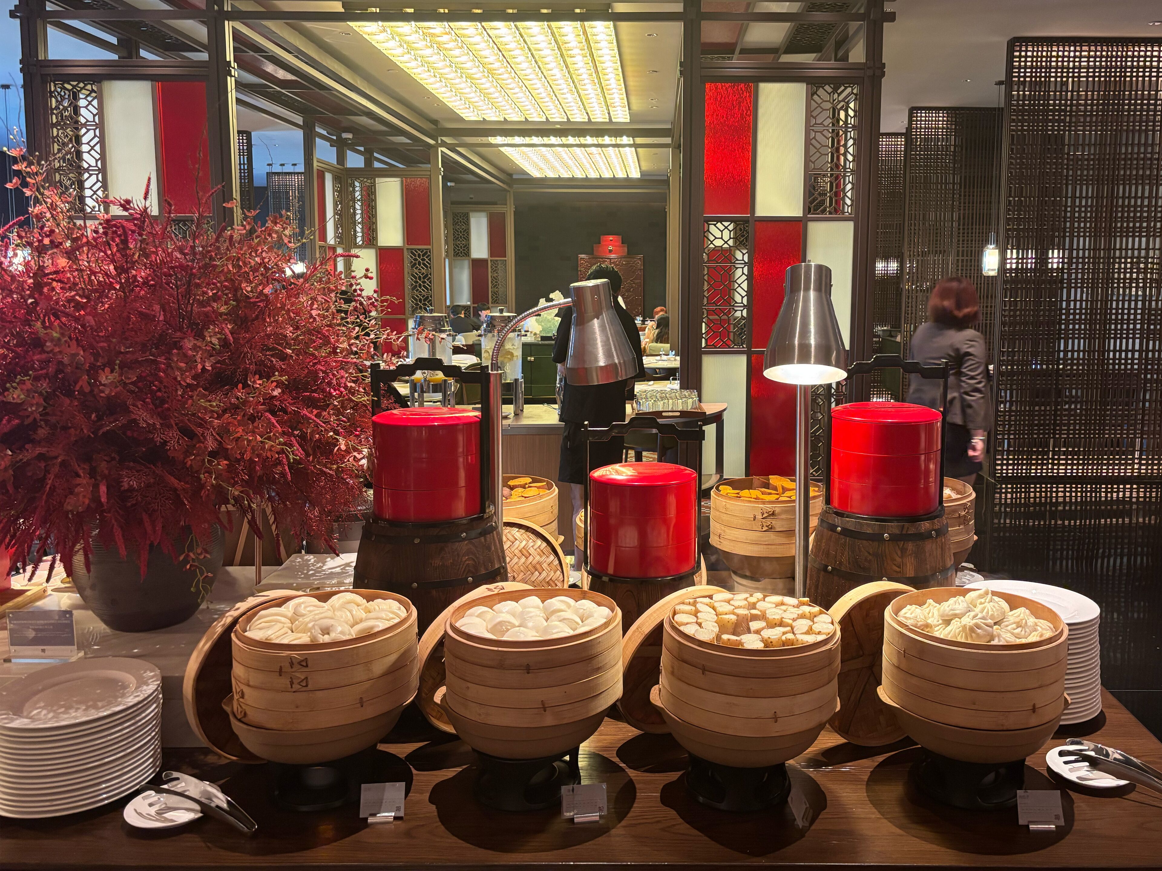 Un buffet avec plusieurs présentoirs à étages remplis de plats chauds, une grande plante en pot et un intérieur moderne aux couleurs rouge et blanche.