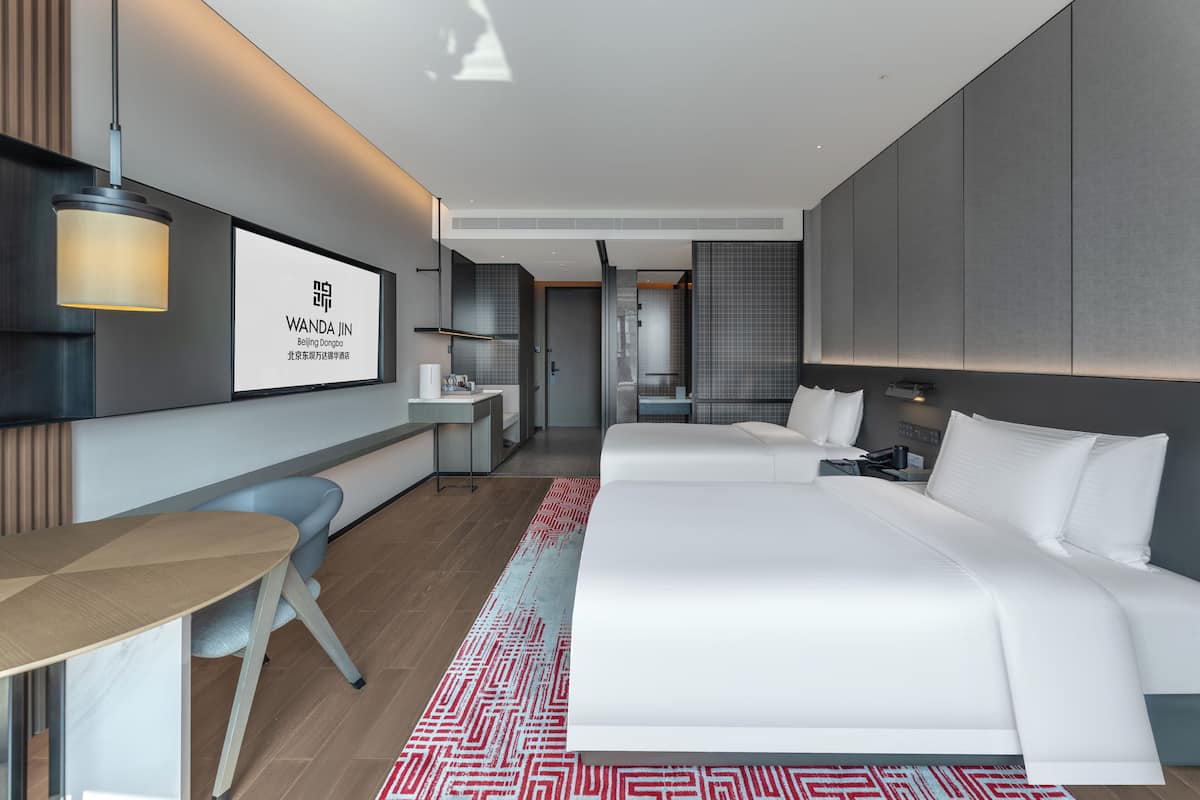 A modern hotel room with a large bed, a desk, a chair, and a TV screen displaying the hotel's name.