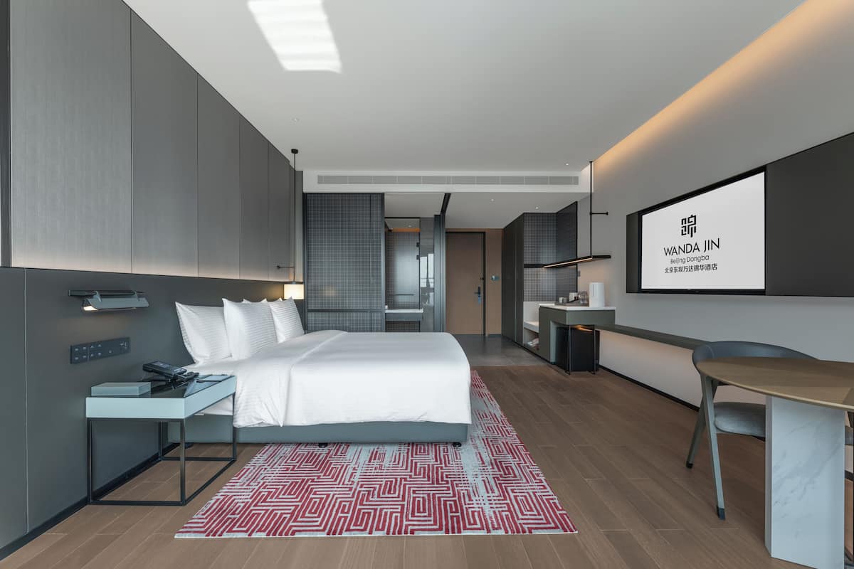 A modern hotel room with a bed, bedside table, desk, and a TV screen displaying the hotel's name.