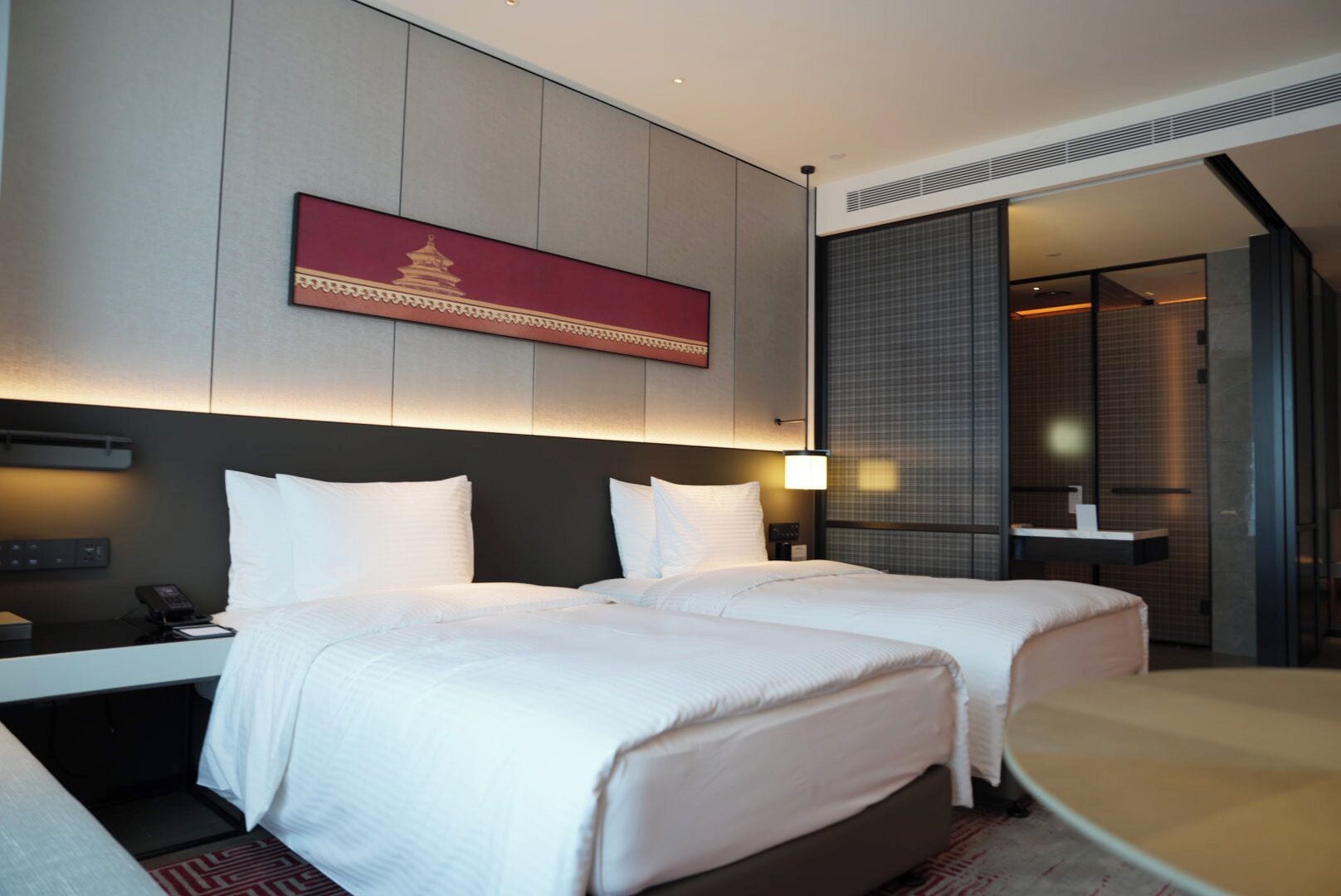 Une chambre d’hôtel avec deux lits, un bureau avec un téléphone, une œuvre d’art accrochée au mur et une salle de bain visible à travers une porte en verre.