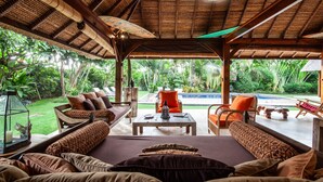 A covered outdoor seating area with a pool view, wooden furniture, and a thatched roof.