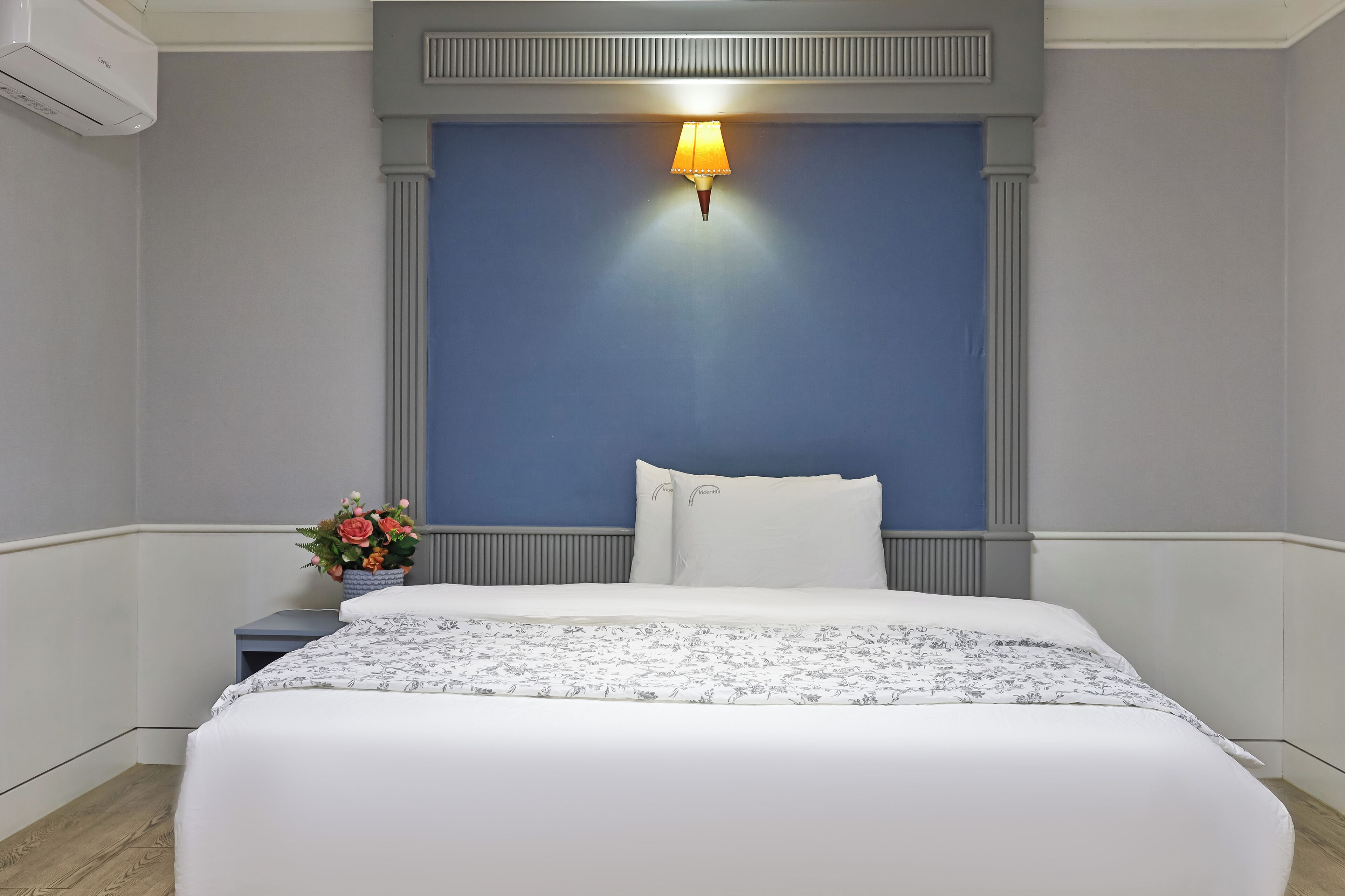 Une chambre d’hôtel avec un grand lit, une table de chevet ornée d’un vase de fleurs et une lampe fixée au mur.