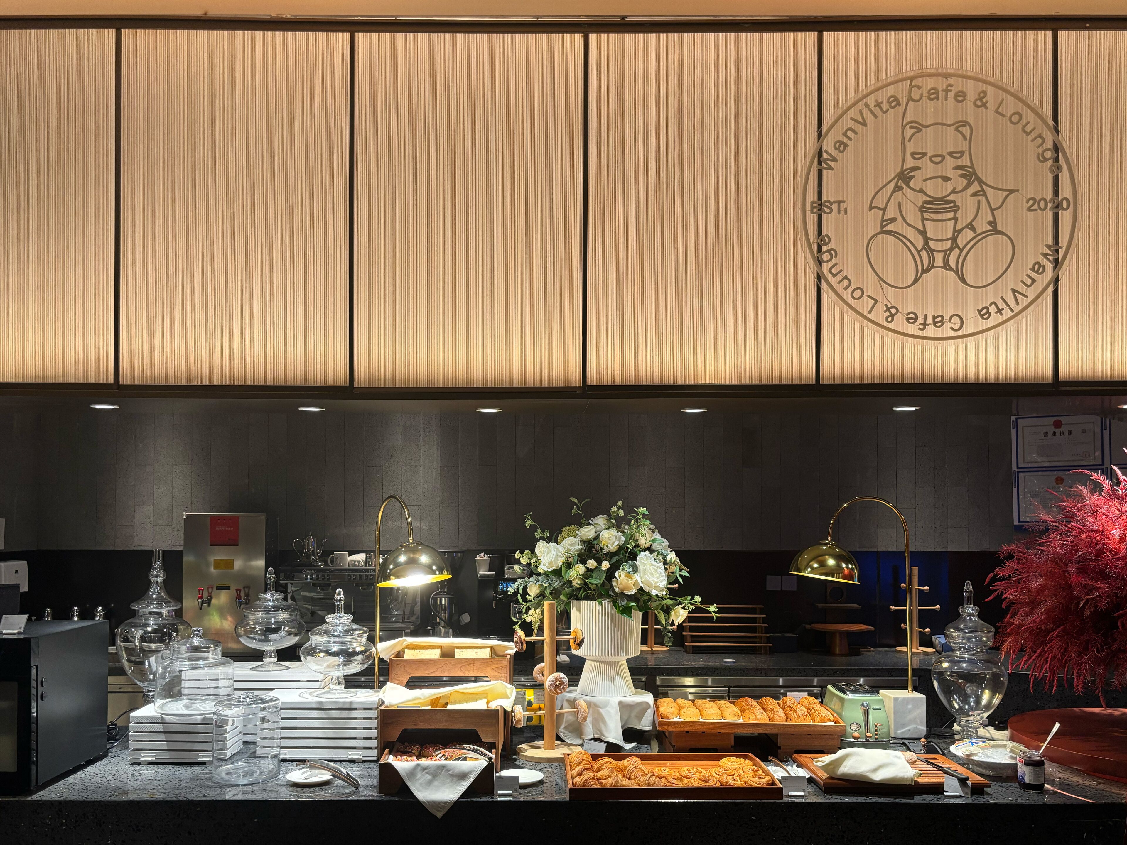 A buffet setup with various food items and glassware, a decorative vase with flowers, and a logo on the wall.