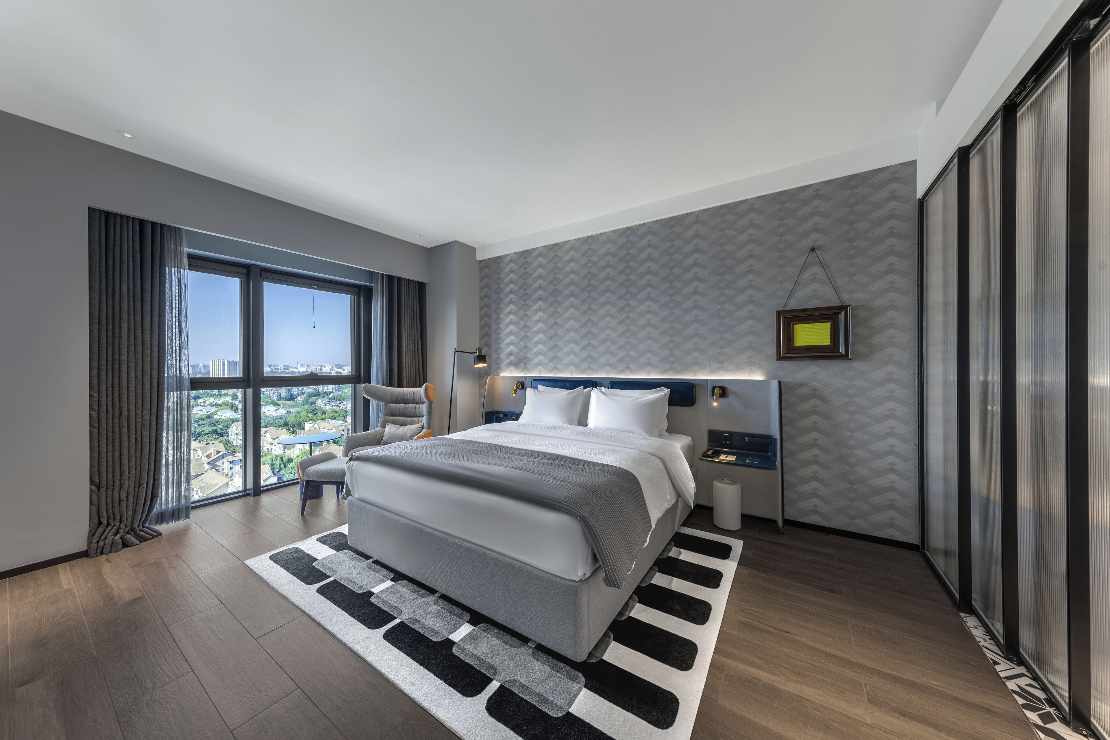 A modern hotel room with a large bed, a city view, a grey patterned wall, and a black and white rug.