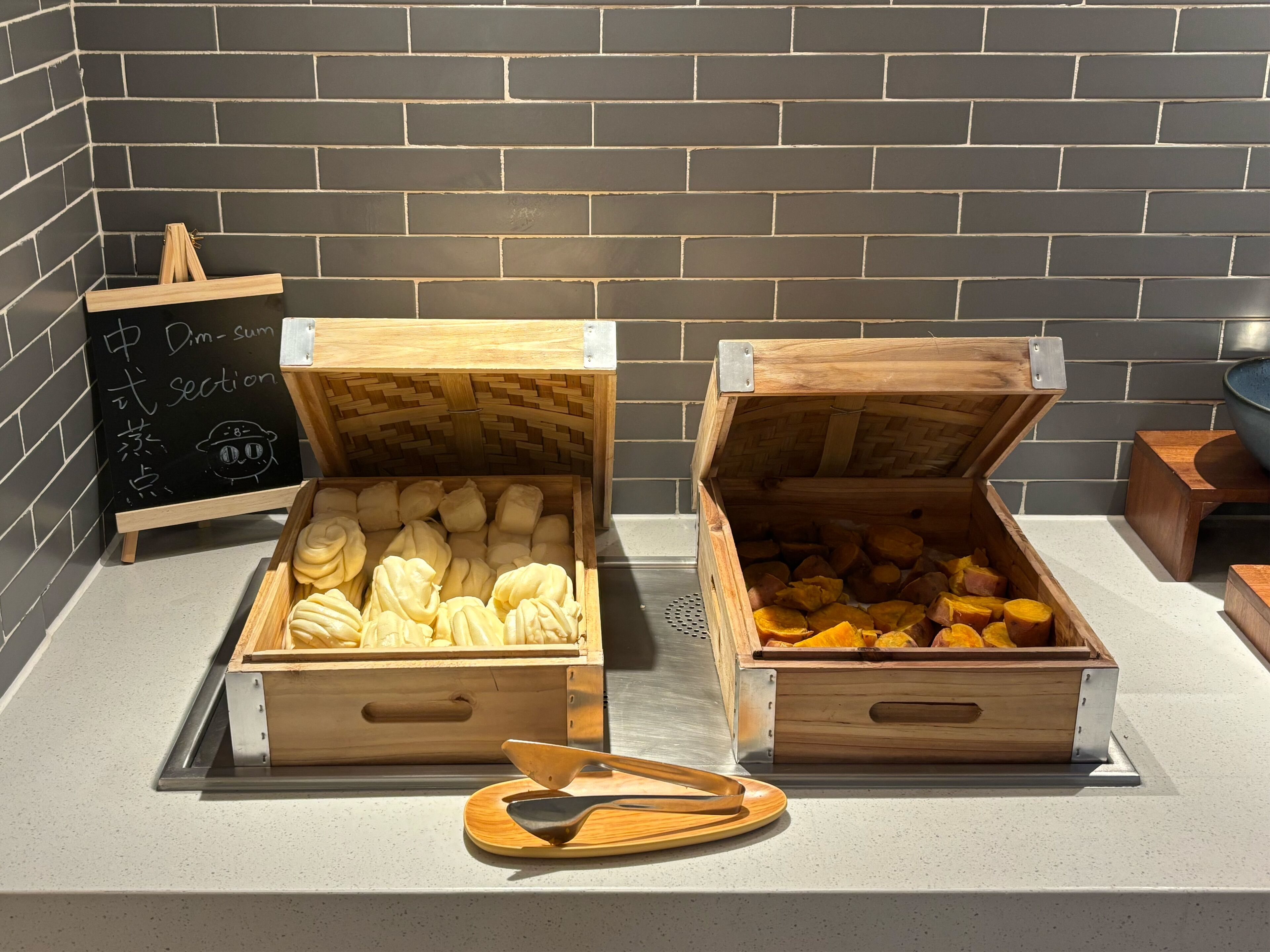 Dos cofres de madera con tapas abiertas que contienen pasteles y batatas, una pizarra con escritura y una cuchara de madera sobre una encimera.