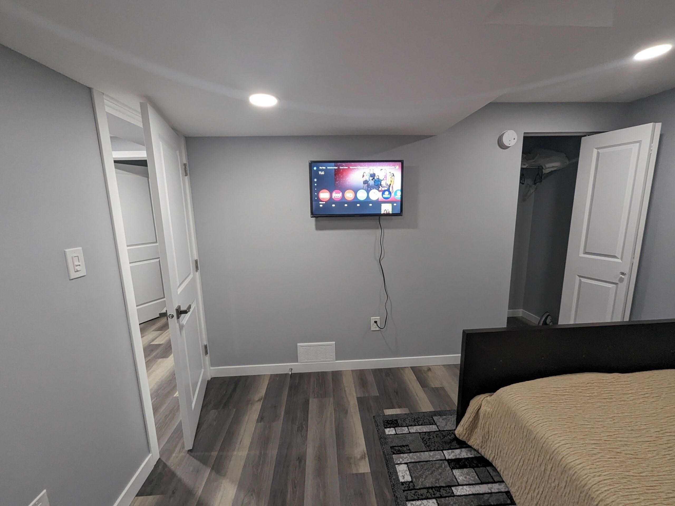 A bedroom with a bed, a TV mounted on the wall, a door, and a vent.