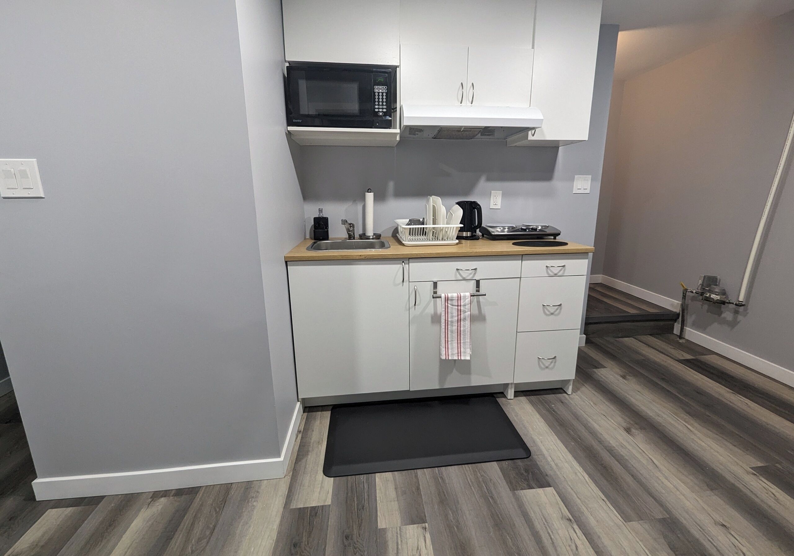 A compact kitchen with white cabinets, a microwave, a sink, and a black mat on the floor.